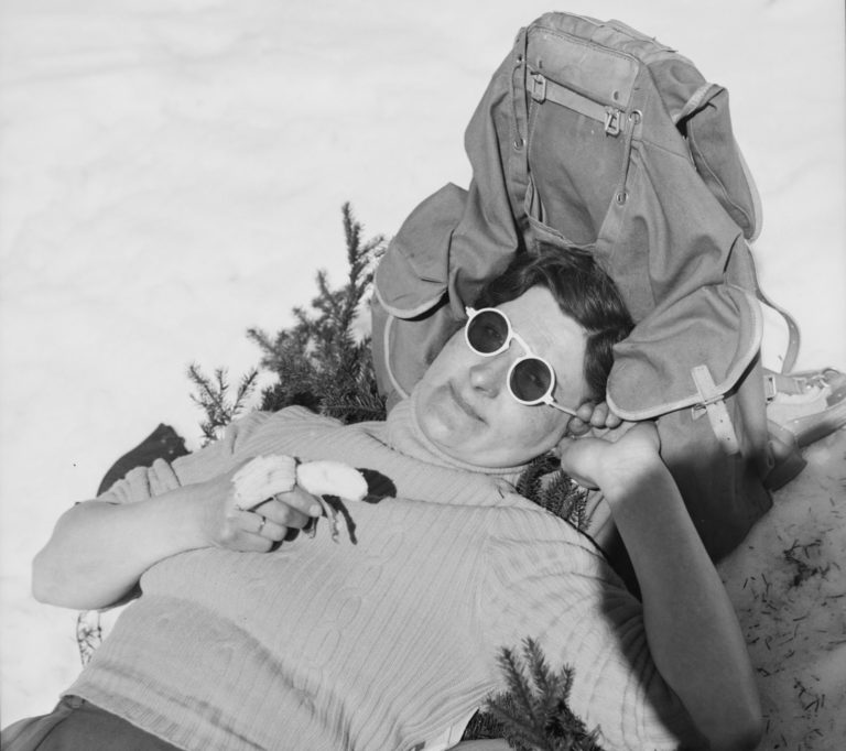 Kvinne med solbriller ligger i snøen, lener hodet mot sekk, spiser banan.