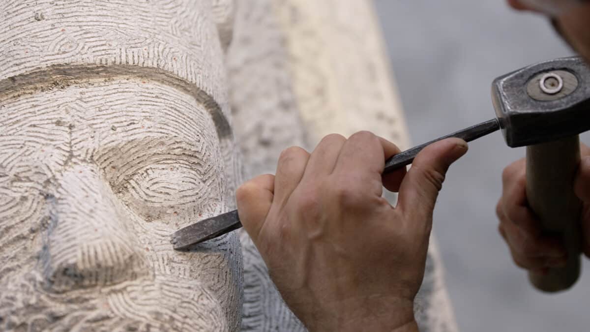 Nærbilde av et par hender som holder hammer og meisel og er i ferd med å hugge ut en kleberstein til en skulptur av Magnus Lagabøte.