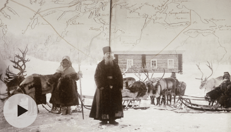 Gammelt fotografi i svart-hvitt som viser samer, reinsdyr og sleder foran en trehytte i et snølandskap.